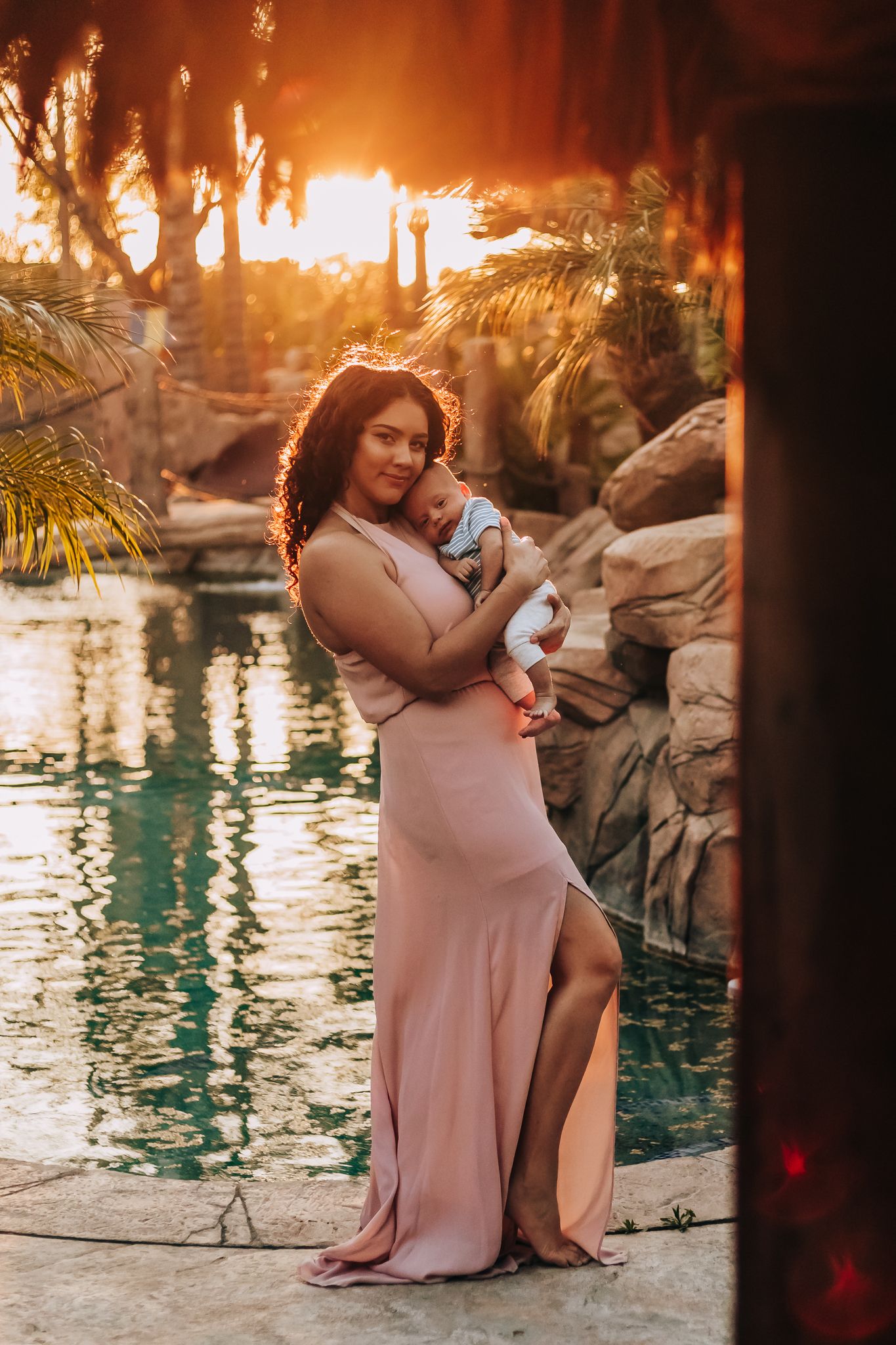 A woman in a blush pink dress holds her baby while standing beside a pool at sunset with palm trees in the background.