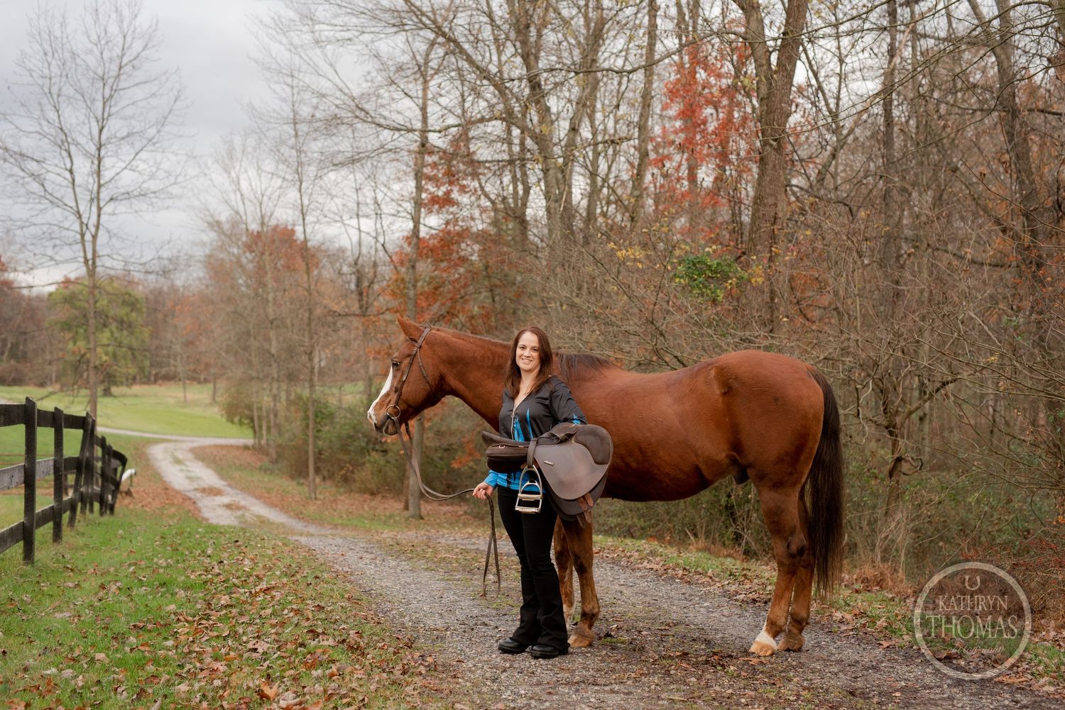 A beautiful chestnut horse and rider pause on an autumn trail with wooden fencing and fall foliage in the background.