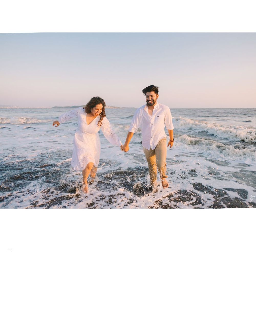Couple playing and splashing in ocean waves at sunset wearing white outfits on the beach.