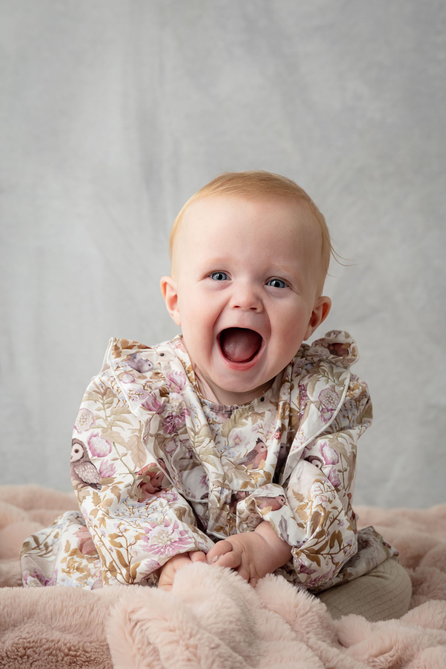 Baby wearing floral print dress smiles joyfully while sitting on pink blanket against grey background.