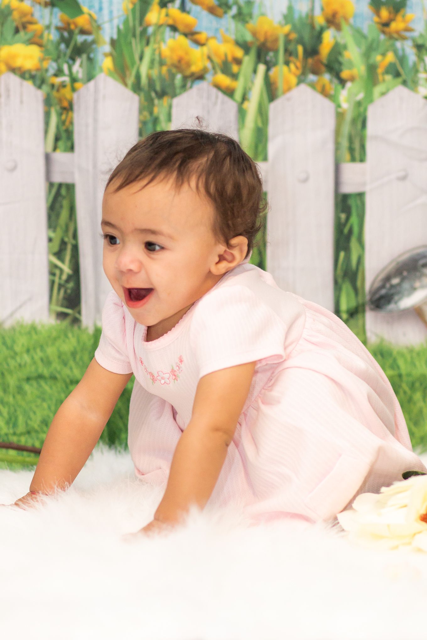 Baby in pink outfit crawls on white surface with yellow flowers and white fence in background.