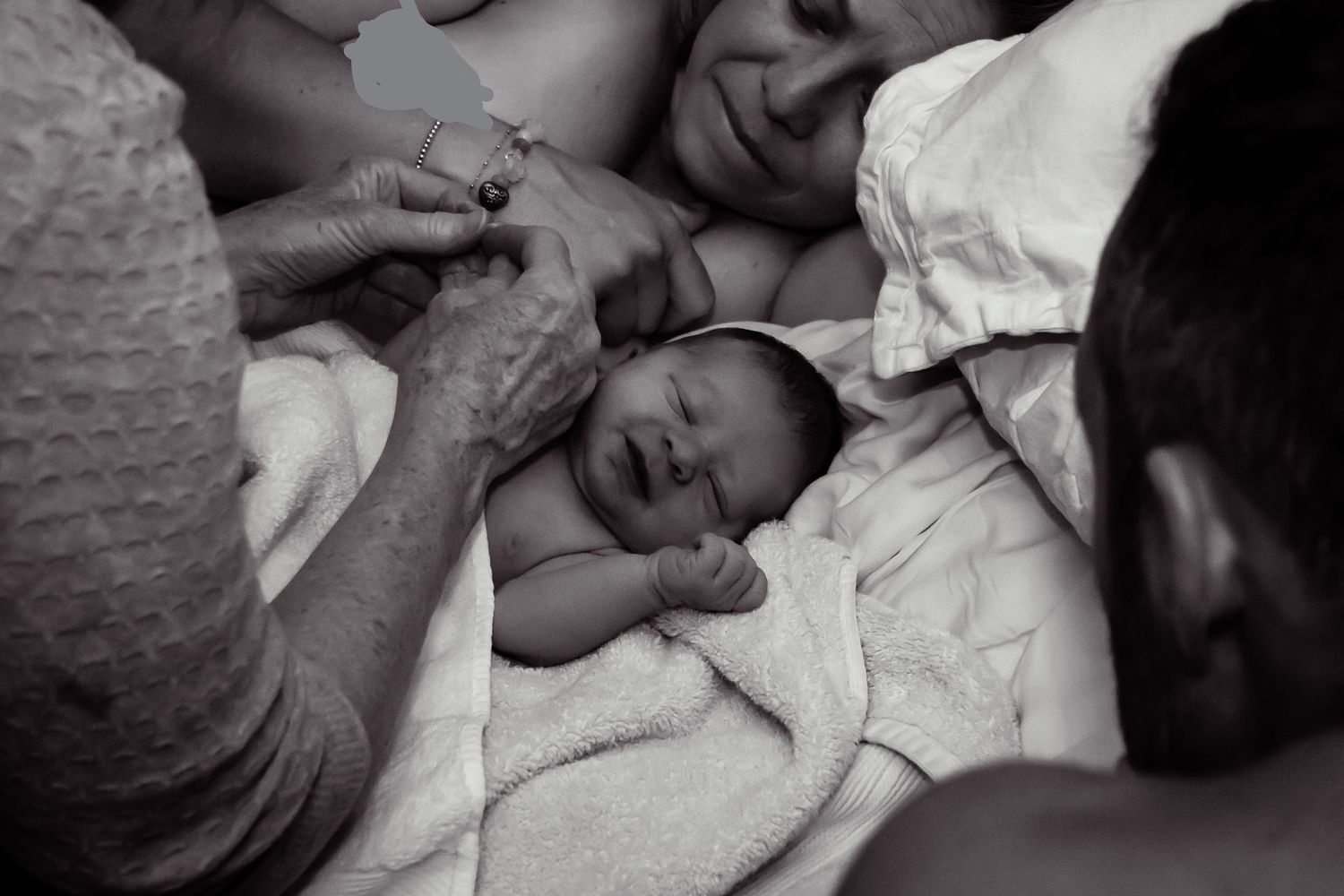 Black and white image of a newborn baby smiling and laying on a bed as a midwife examines her hands.