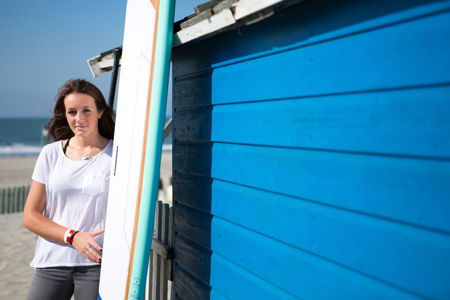 Person standing next to blue beach hut with surfboard on sunny day during business branding photoshoot.