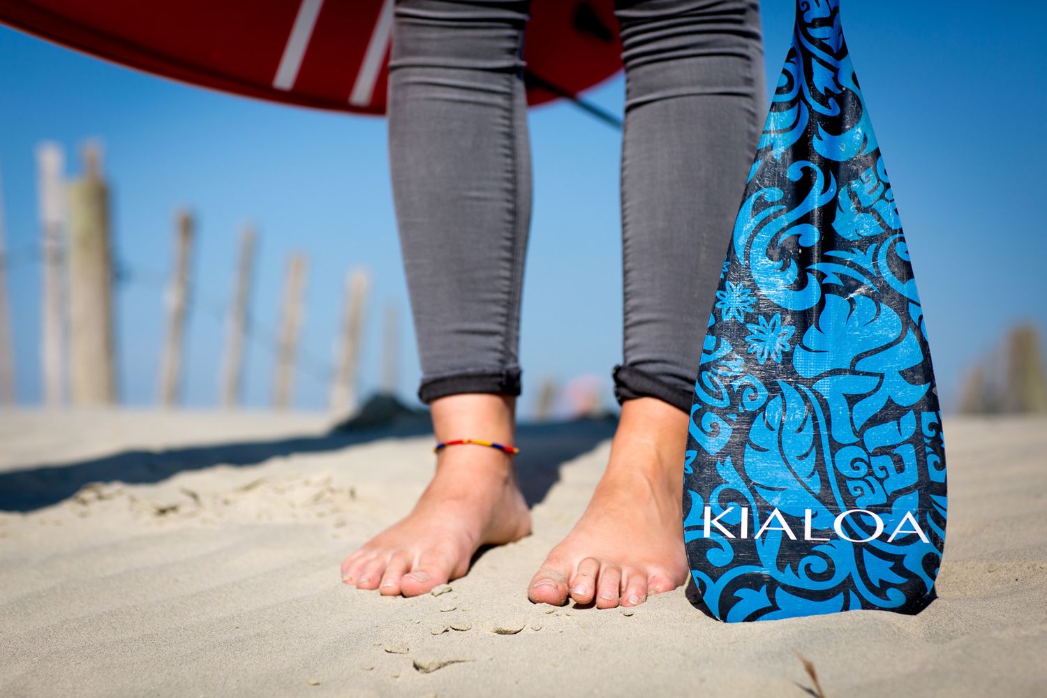 Bare feet in sand next to blue patterned surfboard fin on beach during business branding photoshoot.