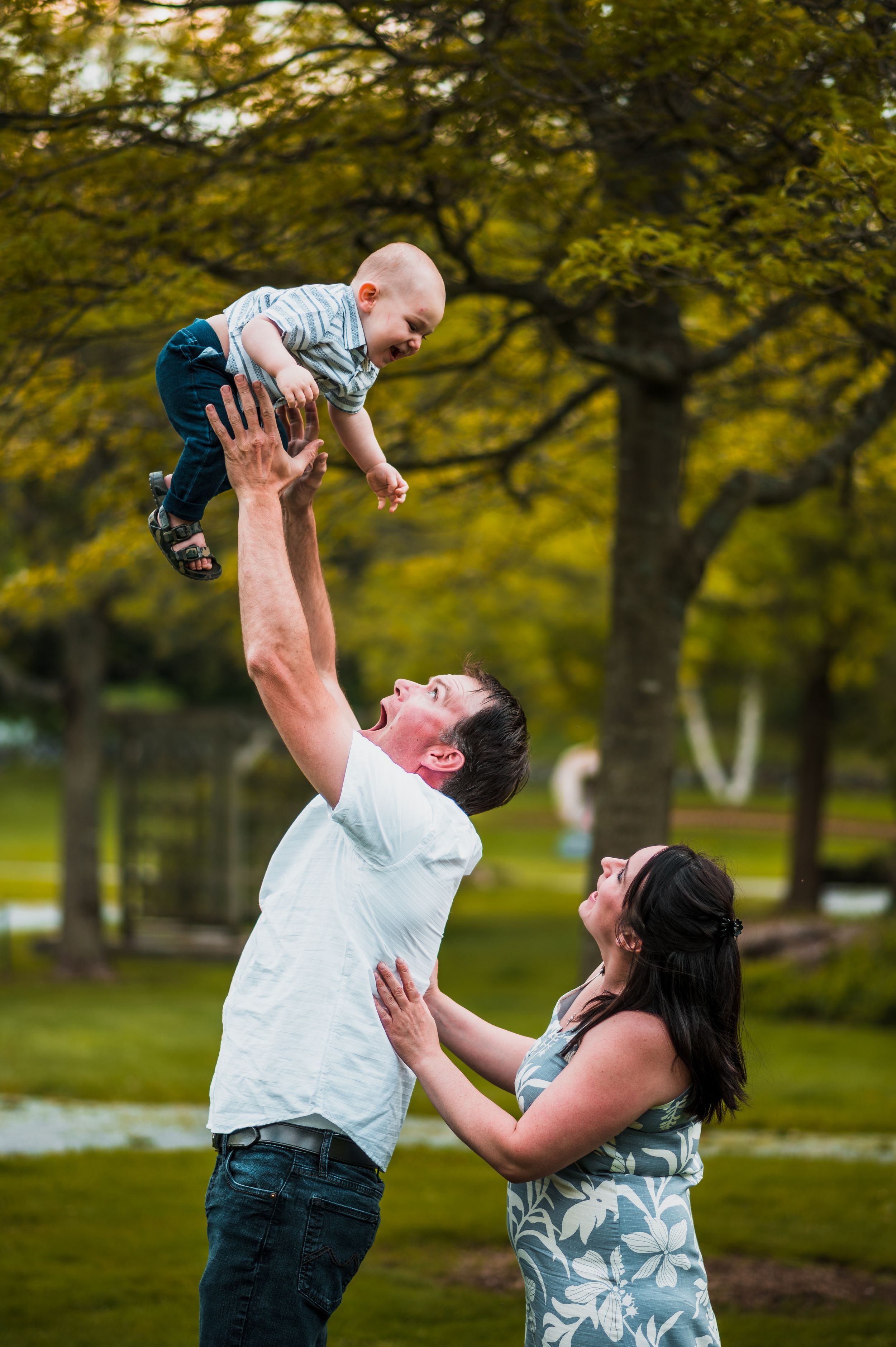 Parents playing with child in a park, lifting them up in the air against autumn trees.