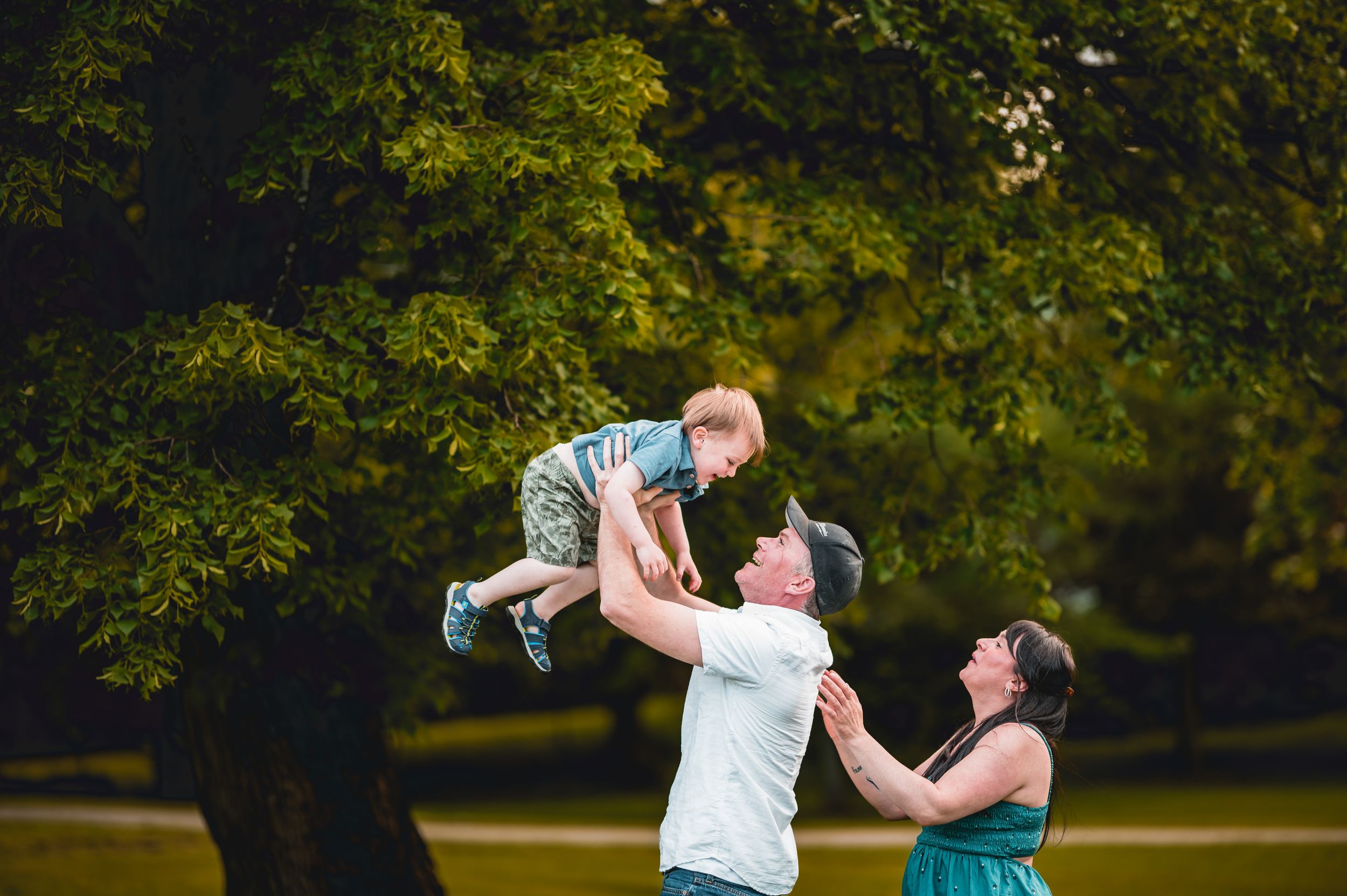 Parents playing and lifting child up during outdoor photoshoot in park setting.