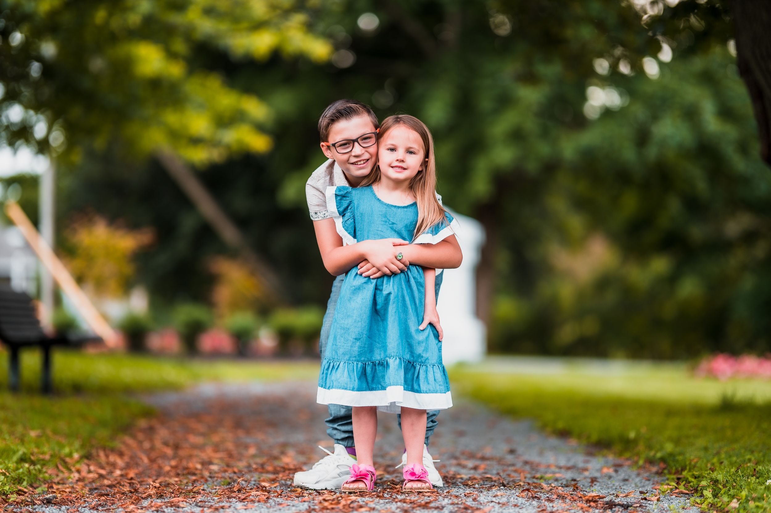 Two children in matching blue dresses play together on a path surrounded by autumn foliage.