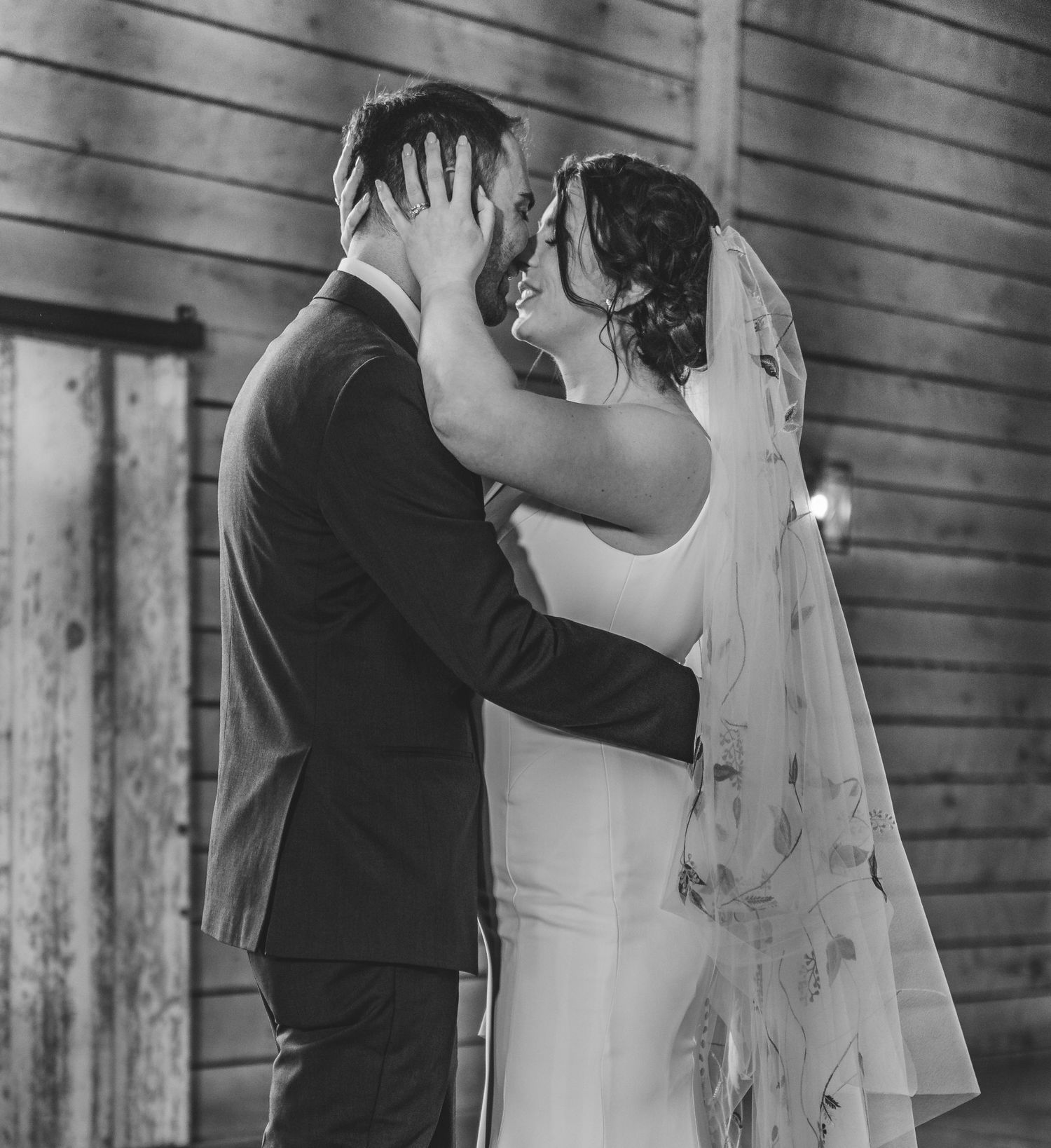 Black and white photo of a romantic wedding moment with bride in flowing veil and groom in suit embracing.