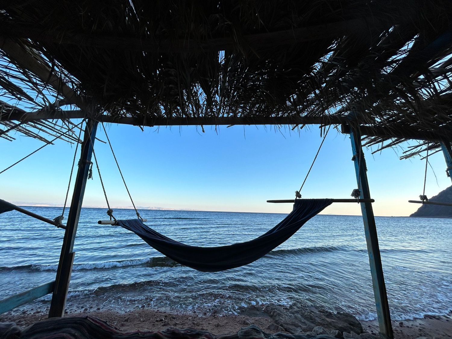 Hammock swings beneath a thatched roof overlooking a serene ocean view on a sunny day.