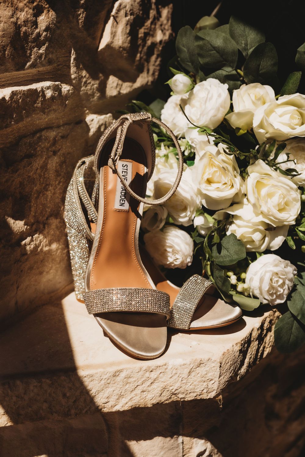Sparkling wedding shoes next to white roses in dramatic lighting.