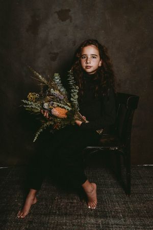 Studio portrait of a red-haired, freckled girl in black, hugging a bouquet of autumn-colored flowers, captured with soft studio light.