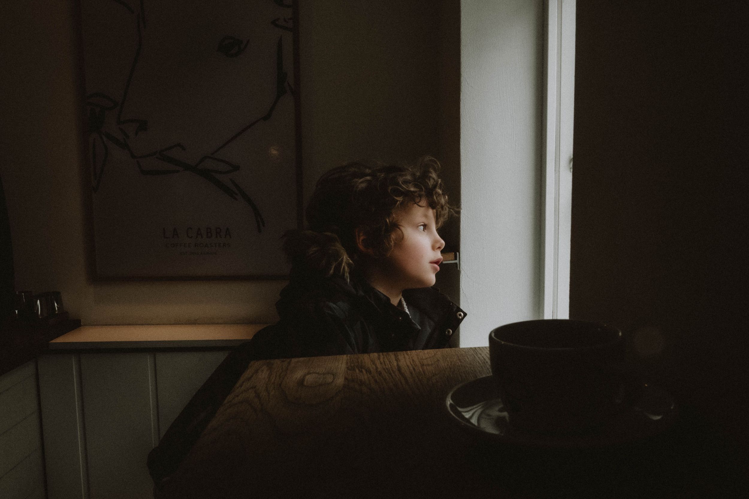 Young child looking out a window in a dimly lit room, illuminated by natural light, preserving a tender and contemplative moment.
