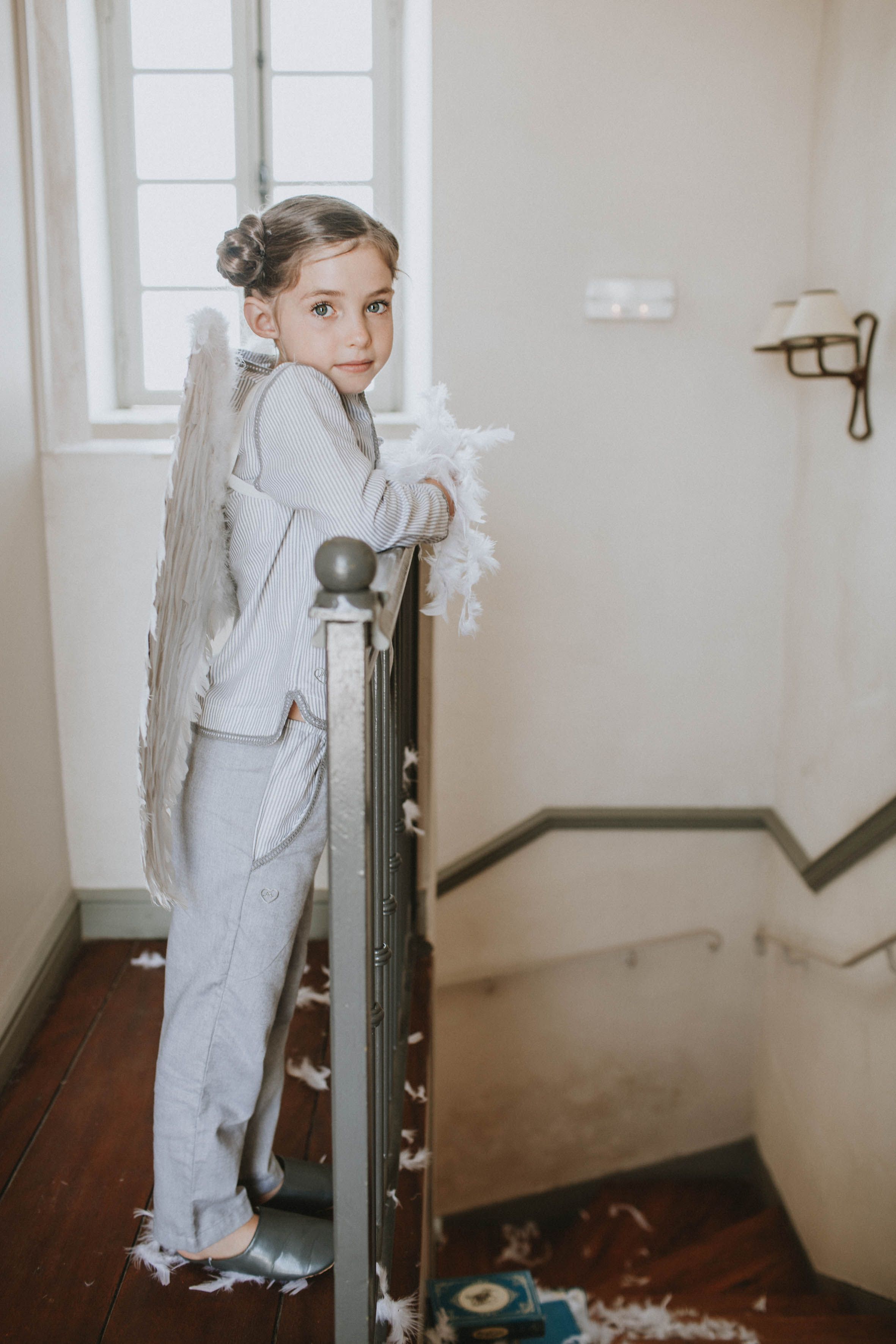 Child in pajamas with angel wings standing at the top of stairs, capturing a dreamy and magical moment in natural light.