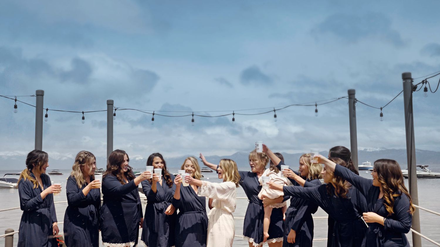 A group in navy and white attire celebrates joyfully together on a beach under string lights and cloudy skies.