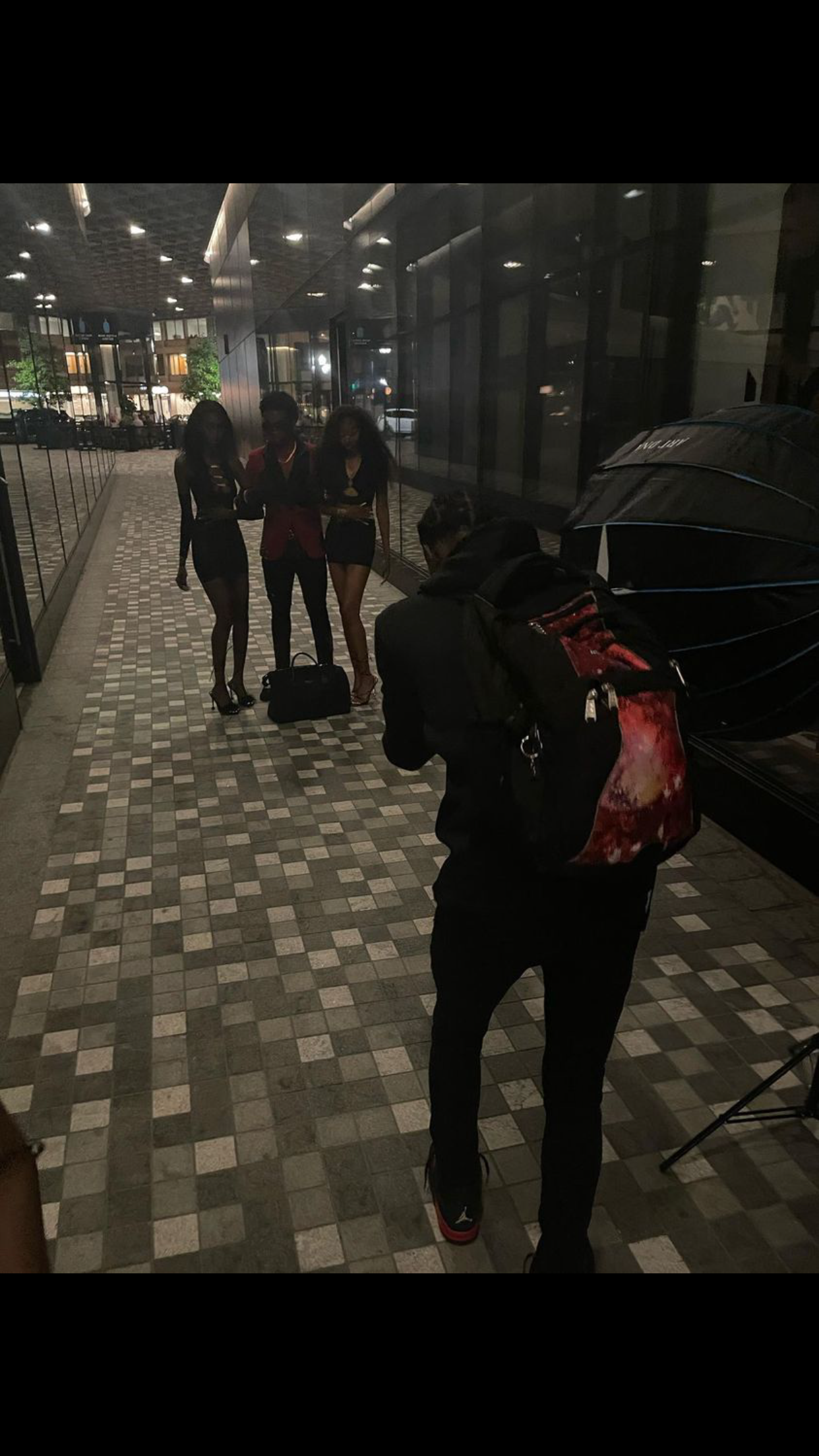 Person with backpack photographing two women posing outside a modern building at night.