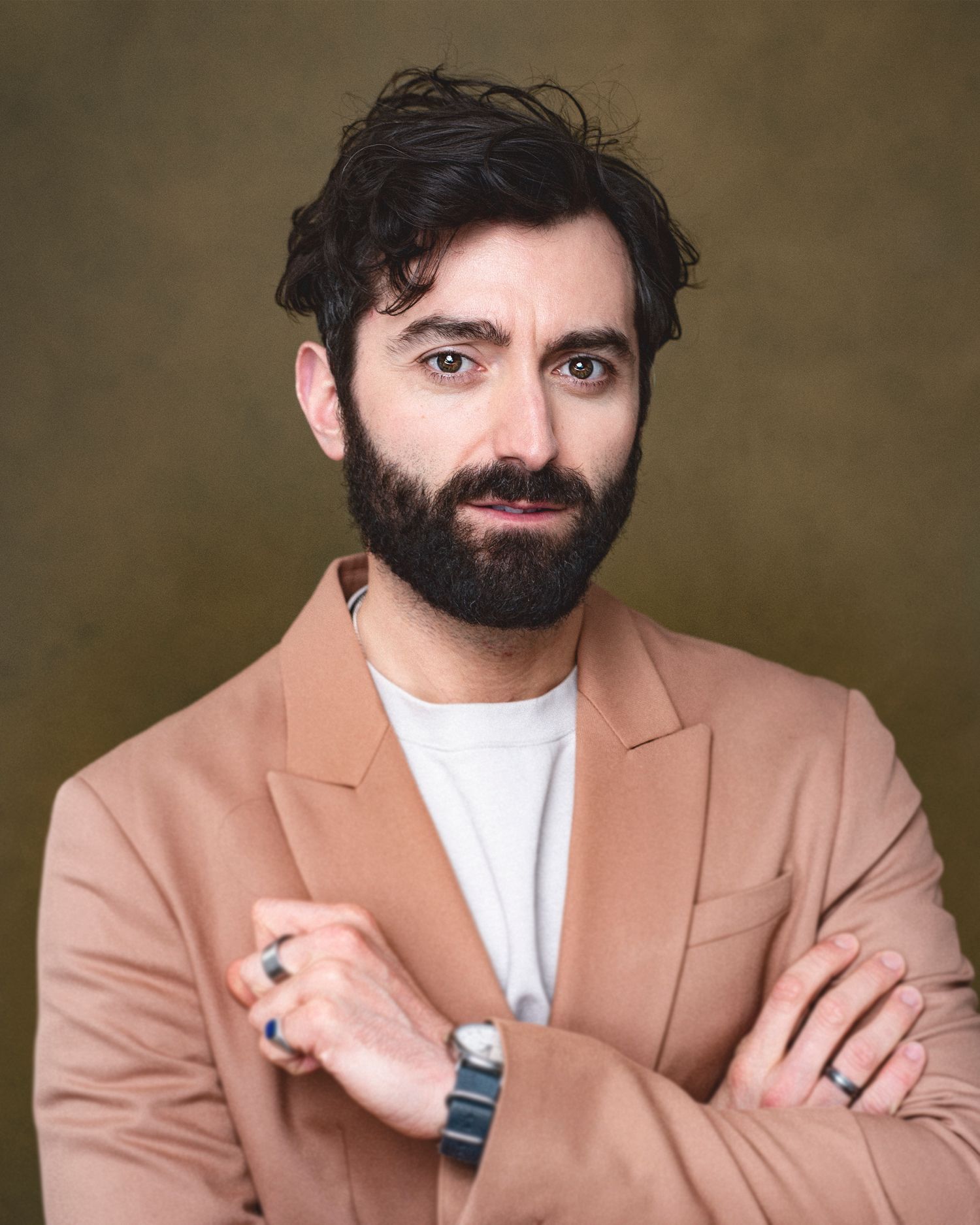 Professional portrait of bearded person in salmon blazer with crossed arms against brown backdrop.