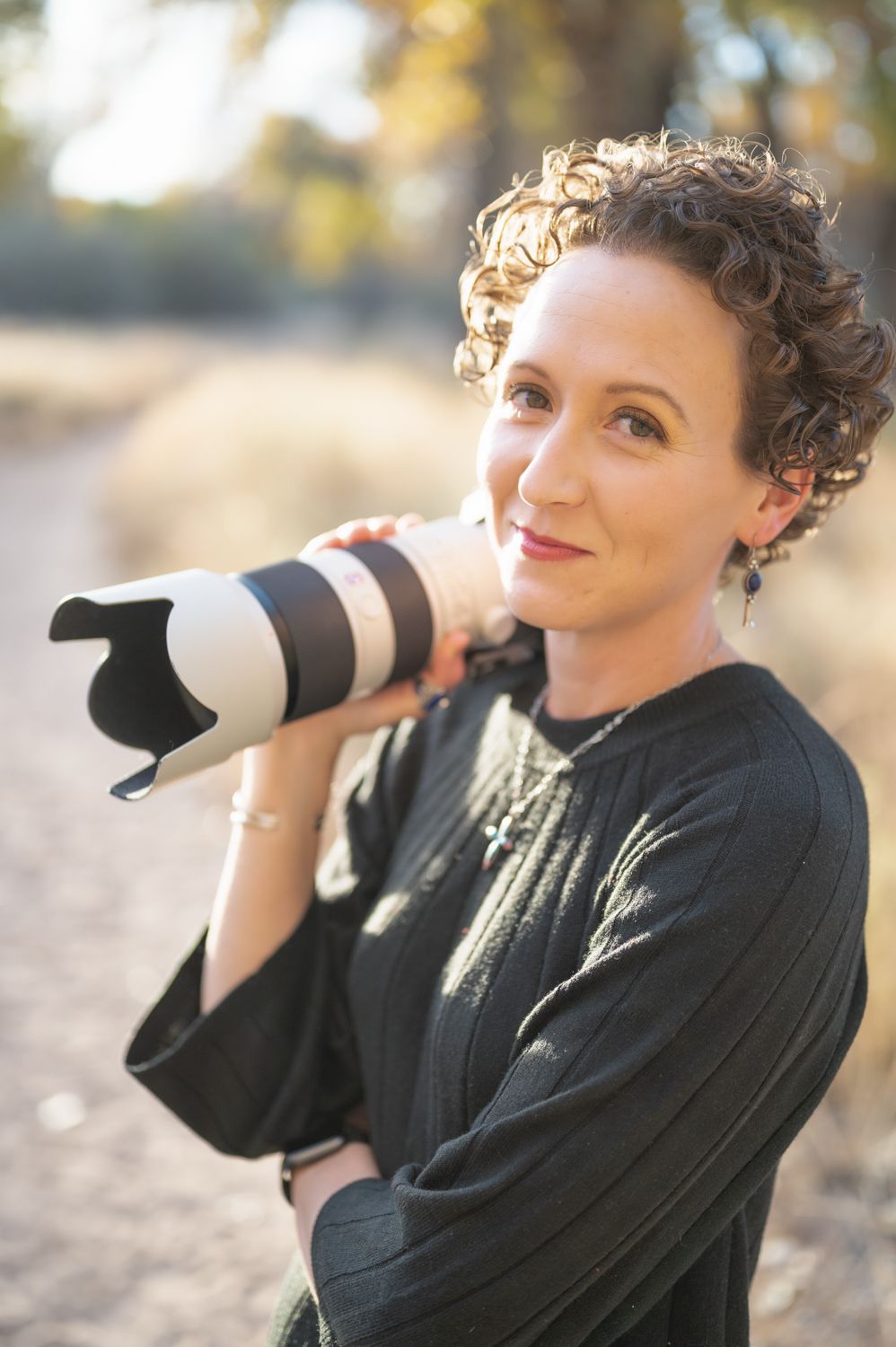 Photographer wearing black top demonstrates using professional camera lens outdoors.