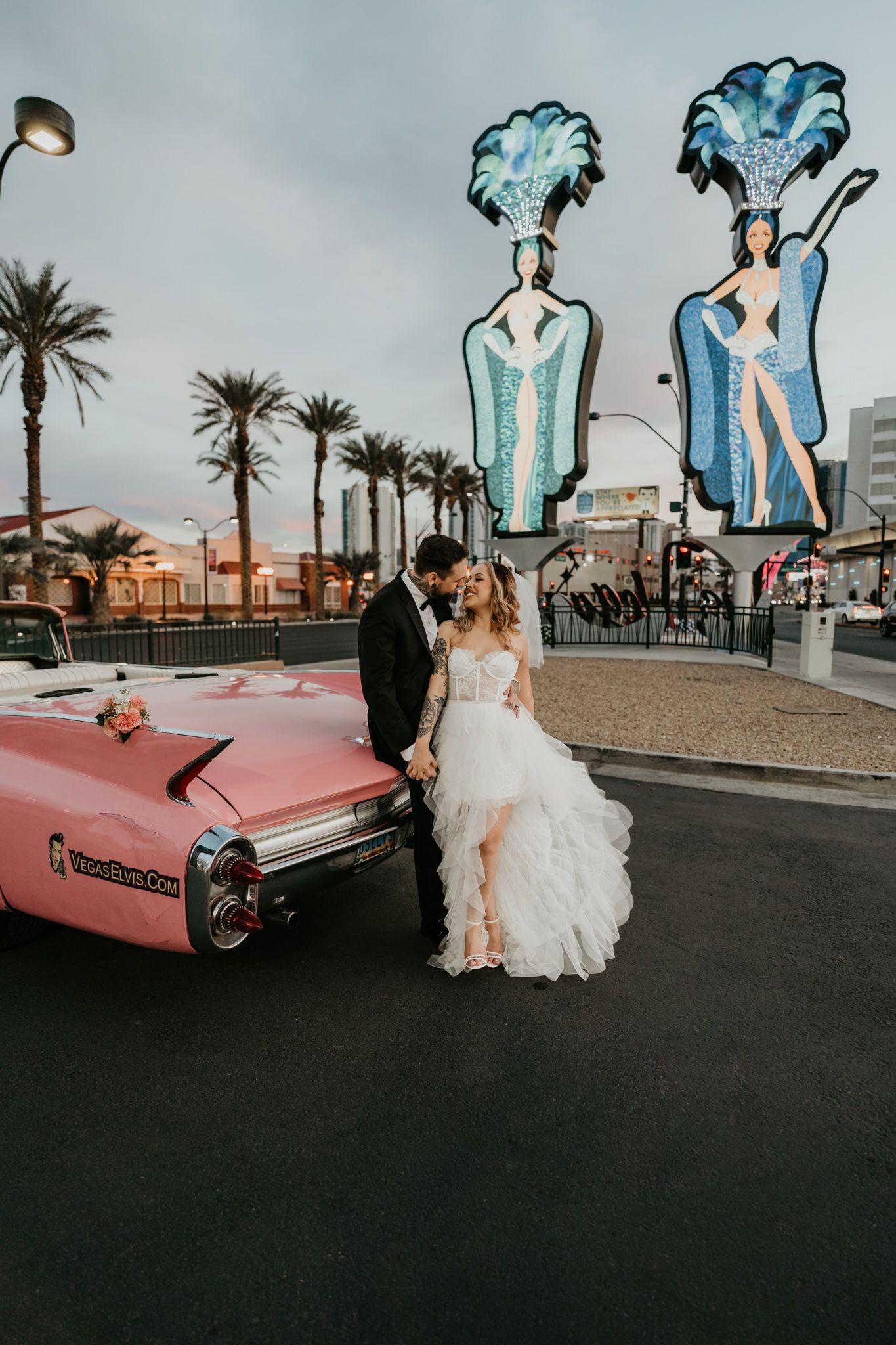 Wedding couple poses with pink vintage car in front of retro Las Vegas signage and palm trees.