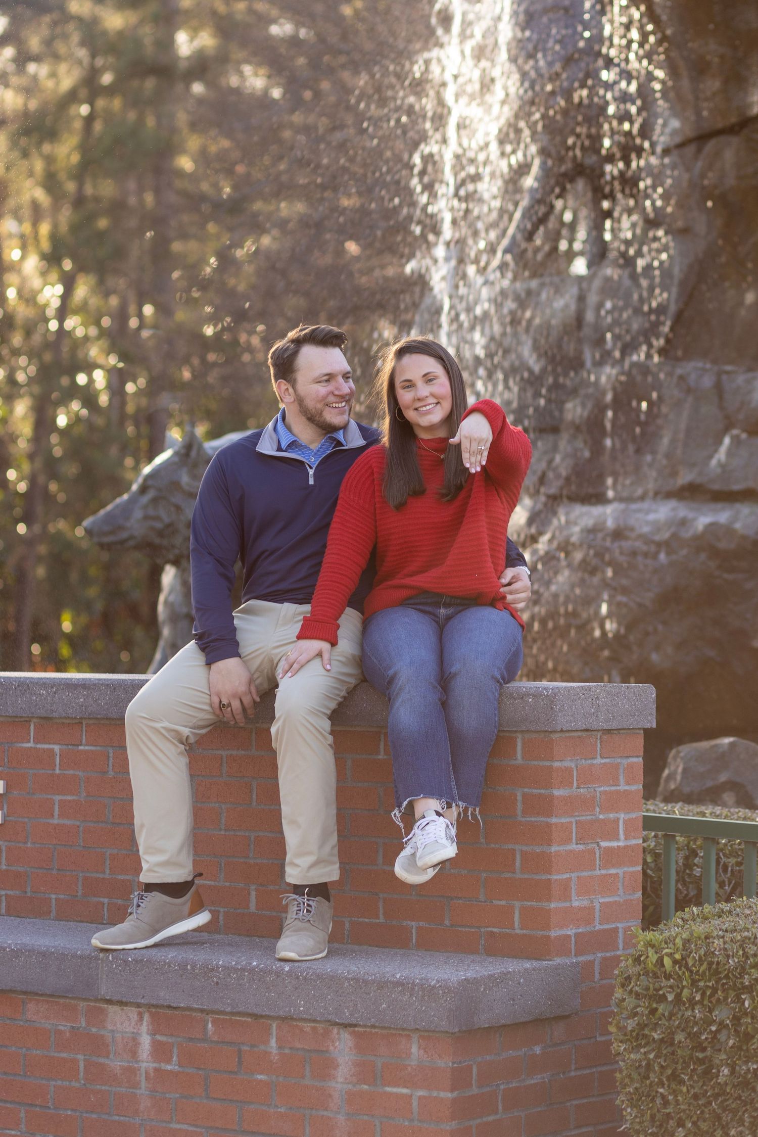 A couple sits in front of a water fountain at their NC State University engagement photoshoot.