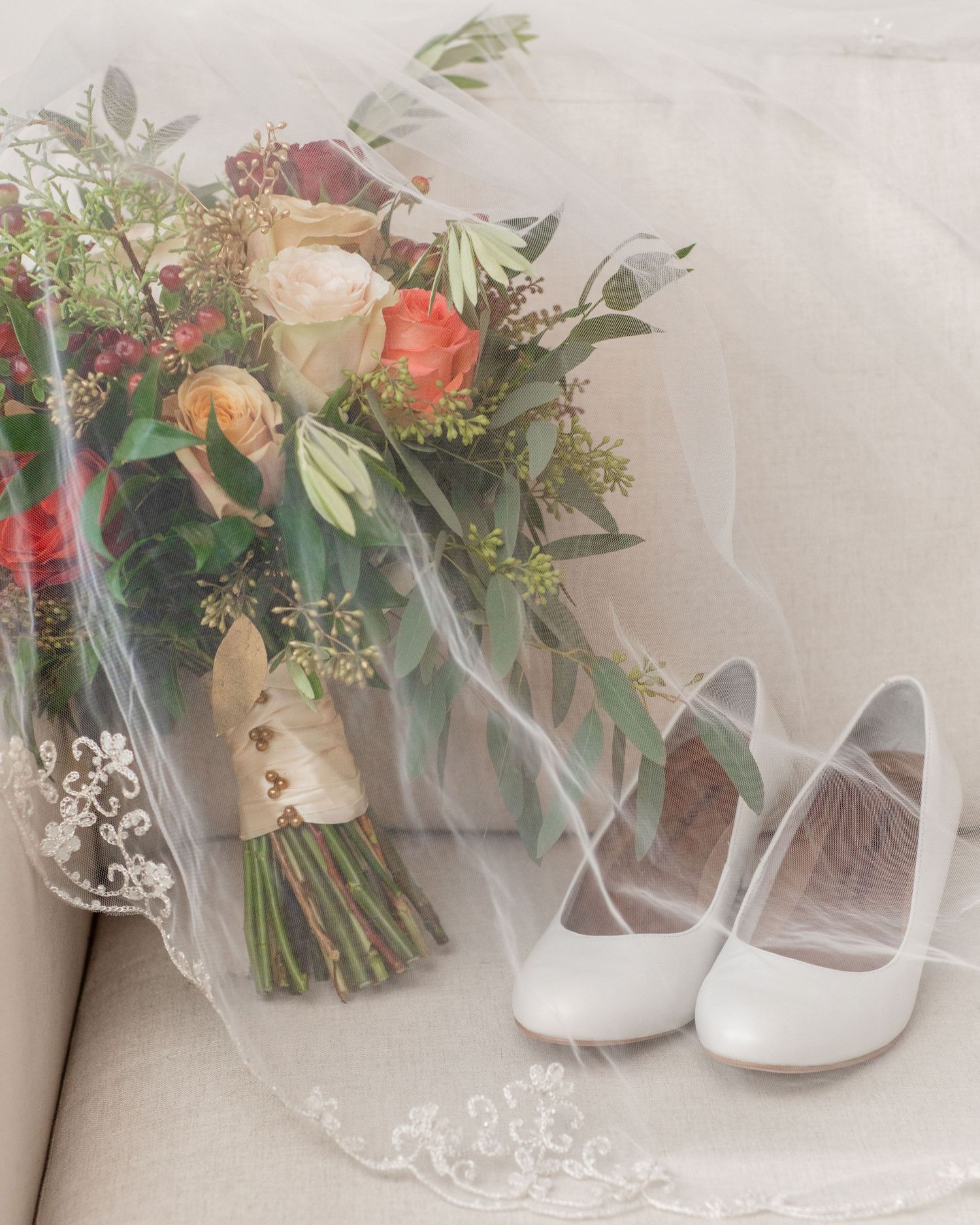 A bridal bouquet with peach and coral roses sits beside white heels on a light surface.