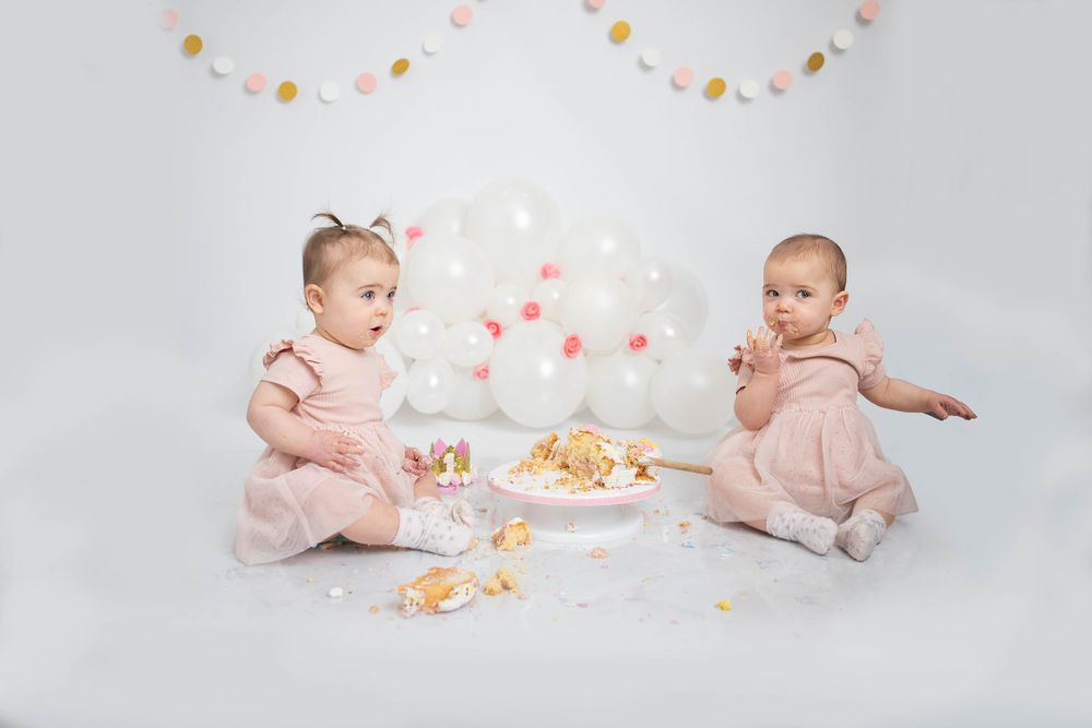 Twin baby girls in pink outfits sitting beside a smashed birthday cake in studio portrait.