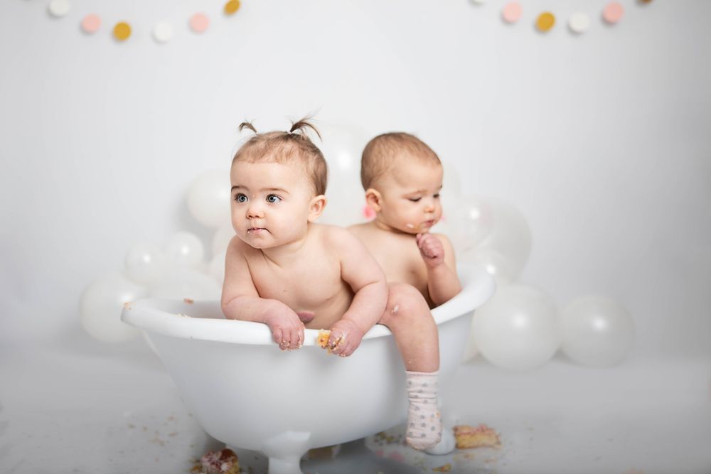 Two babies sitting together in a white bathtub surrounded by balloons in a cake smash session.