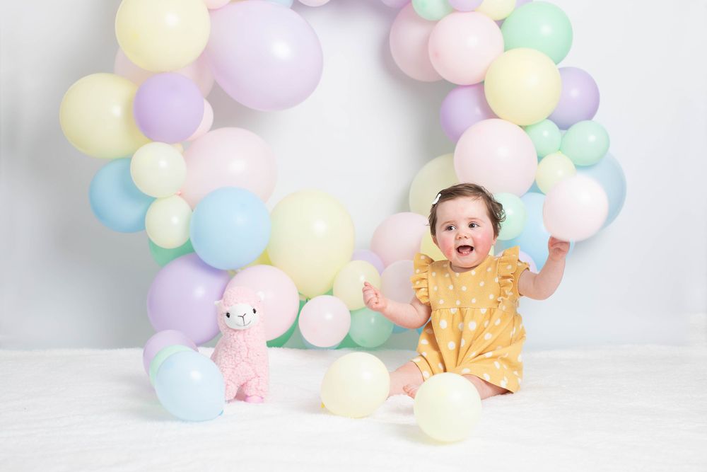 Joyful baby girl in yellow dress with pastel balloon arch and pink llama toy in studio.