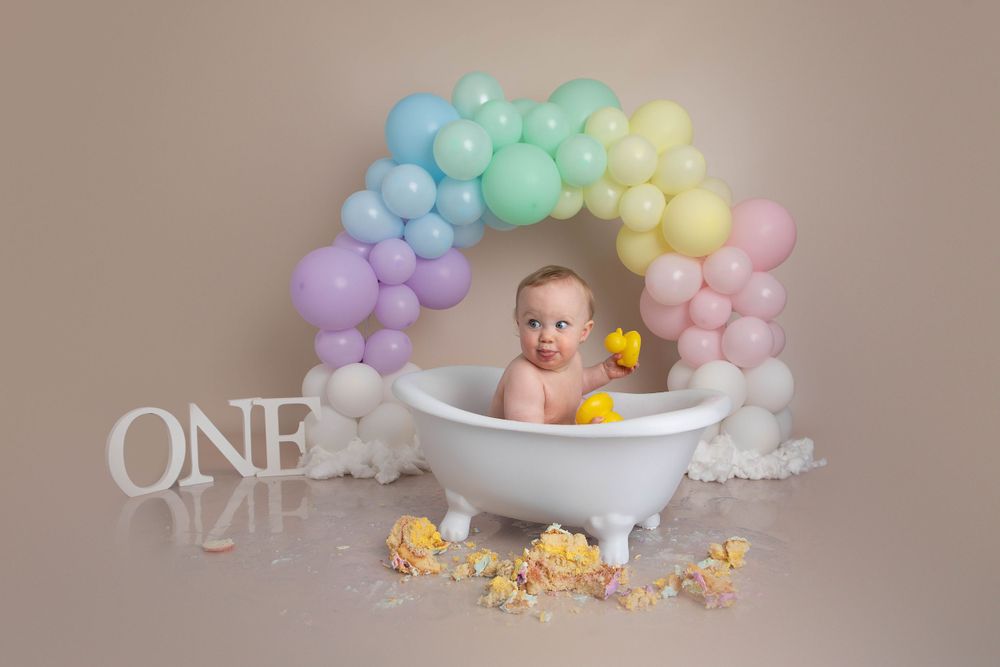 Baby sitting in a mini bathtub with rubber ducks under a pastel rainbow balloon arch for a first birthday.
