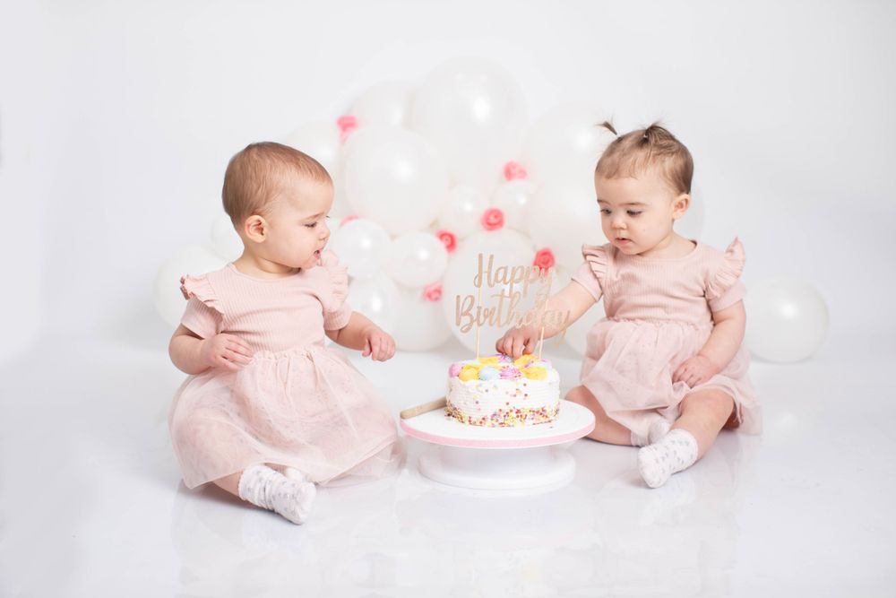 Twin baby girls in pink dresses celebrating their first birthday with a cake and white balloons.