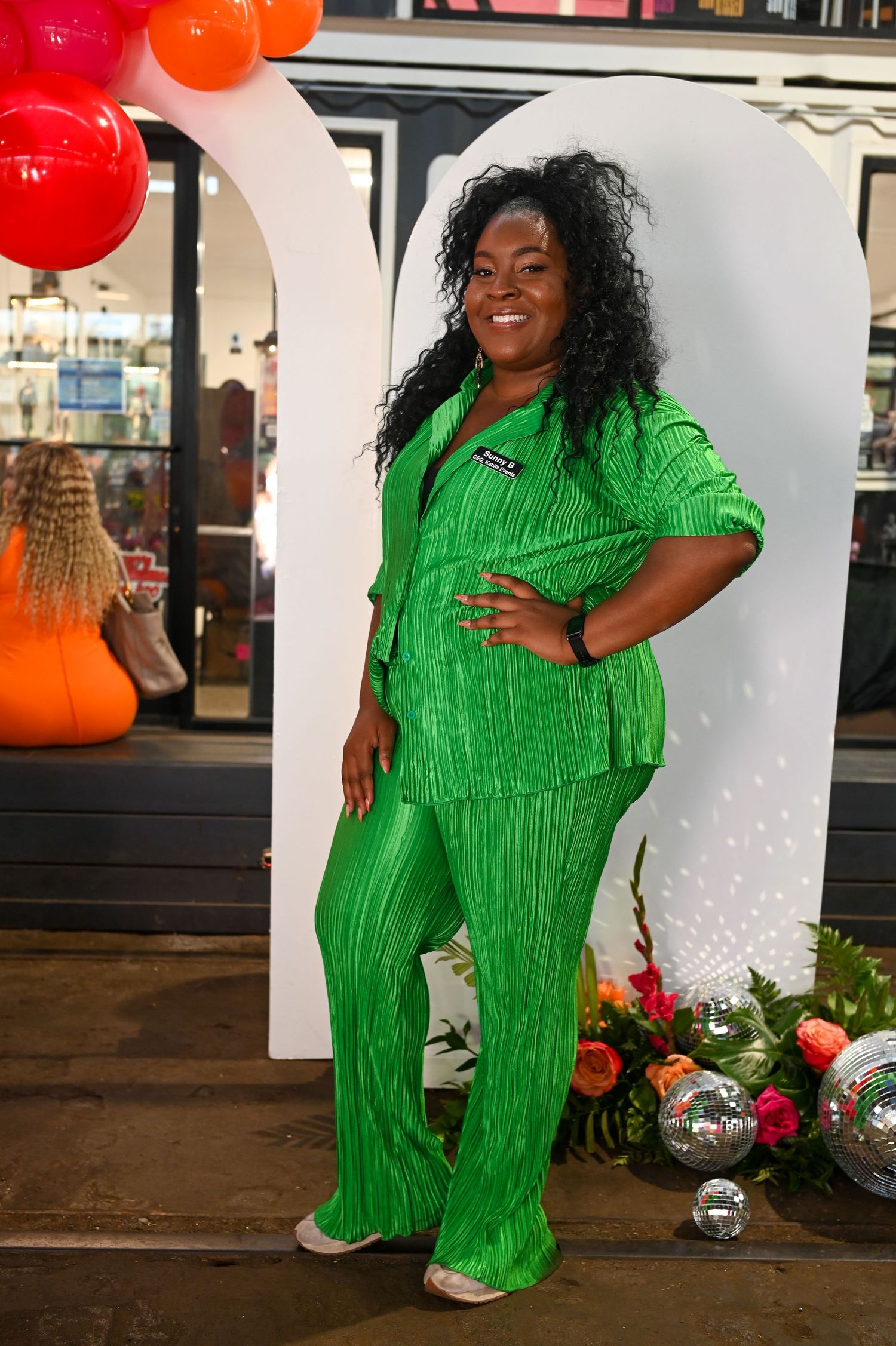 A person wearing a vibrant green textured pantsuit poses against a white backdrop with red balloons.