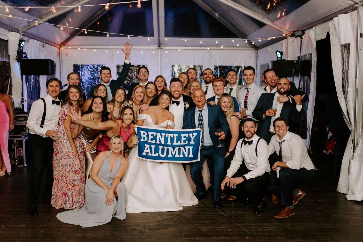Large group photo of wedding guests posing together under an event tent with banners.