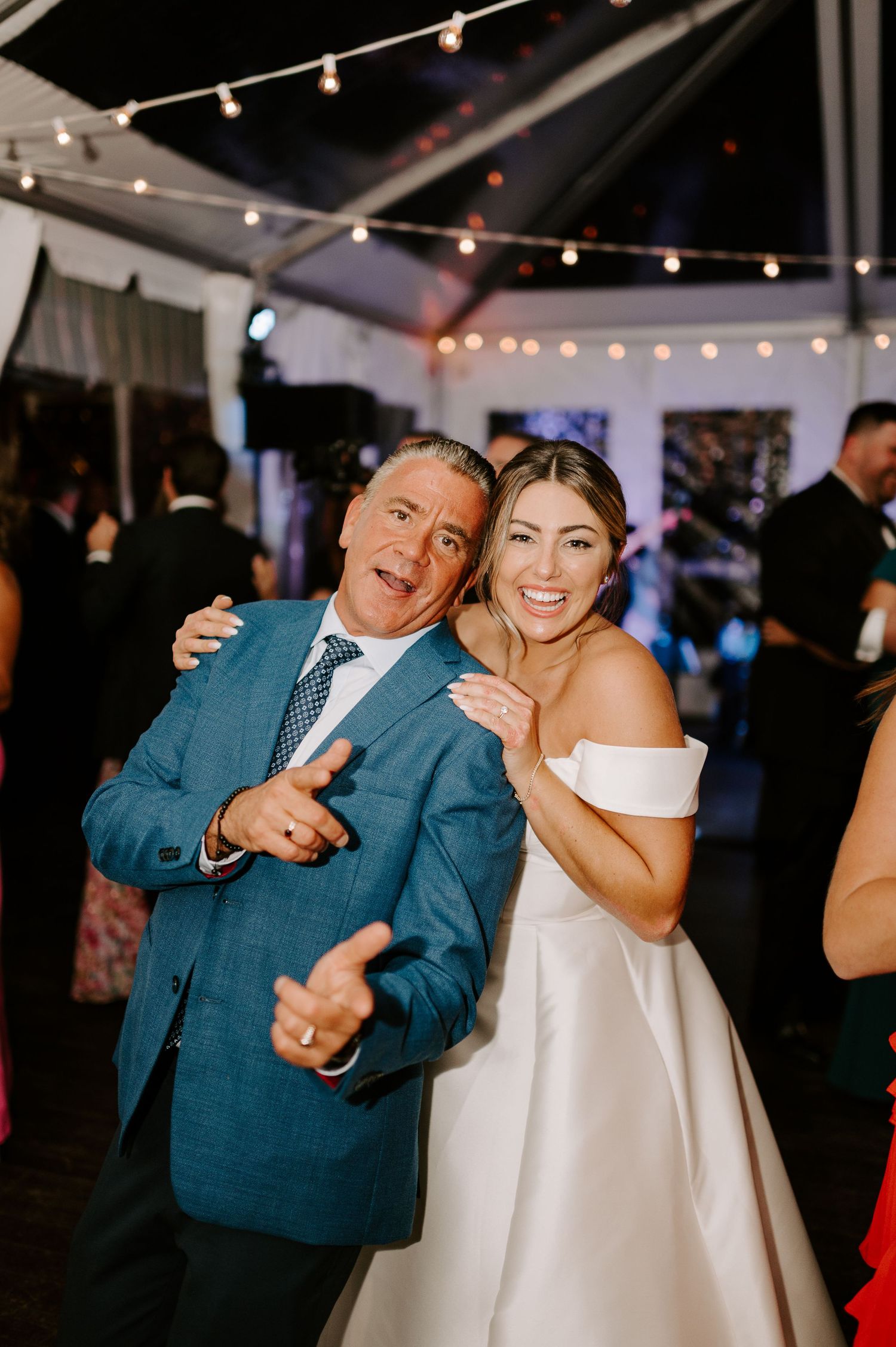 Wedding party members dance and celebrate under string lights at an indoor reception.