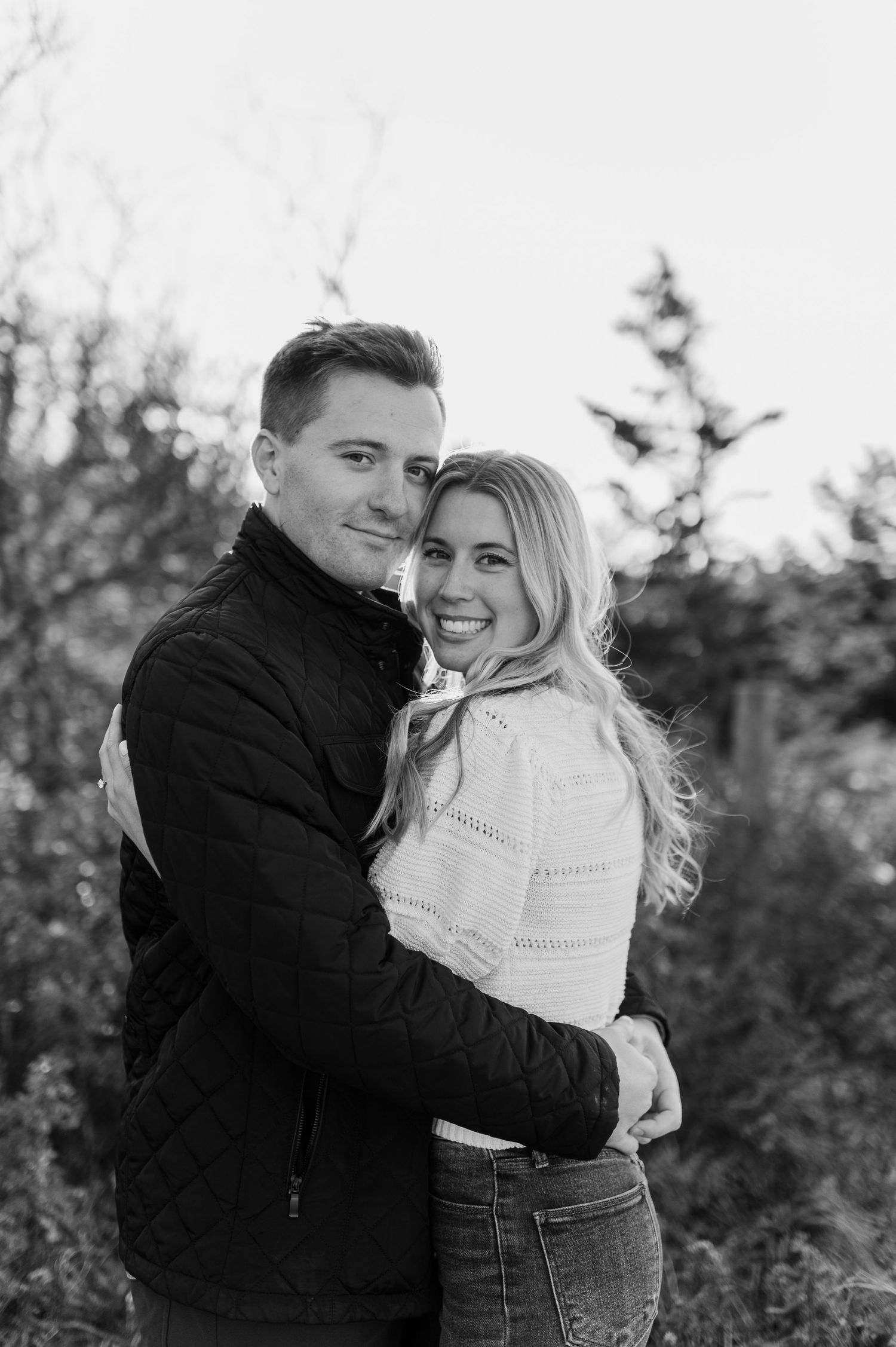 A couple shares warm embraces during an outdoor photoshoot among evergreen trees.