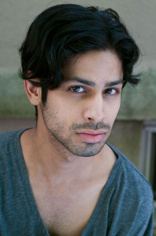Dark-haired man with stubble wearing a gray v-neck shirt, looking away against a blurred background.