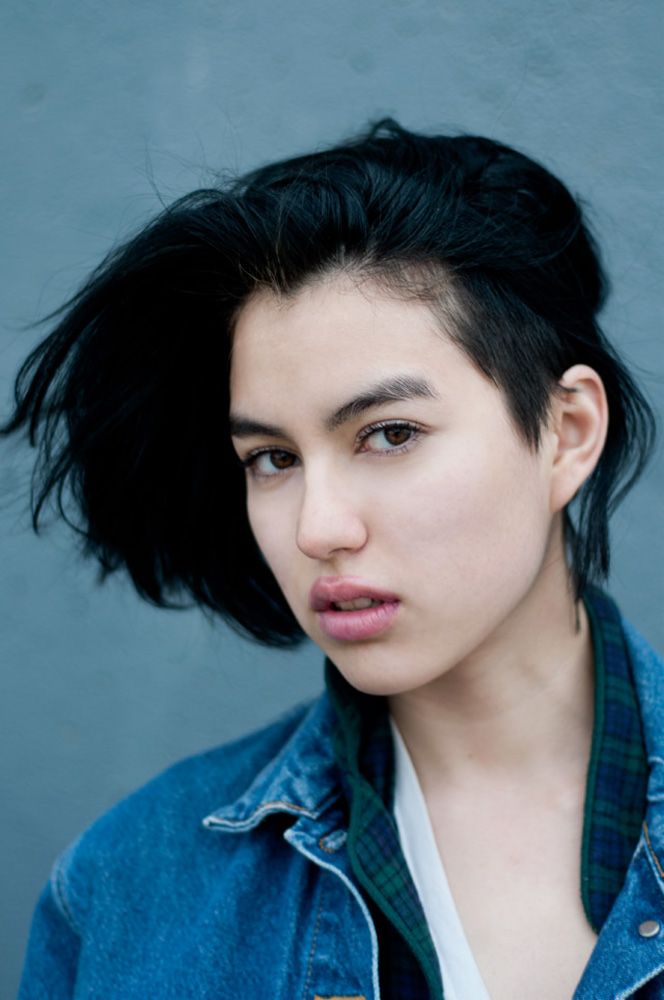 Young woman with black undercut hairstyle wearing a denim jacket against a gray wall background.