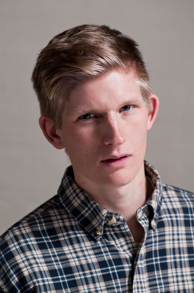 Young man with neat blonde hair wearing a blue and white plaid button-up shirt against a gray background.