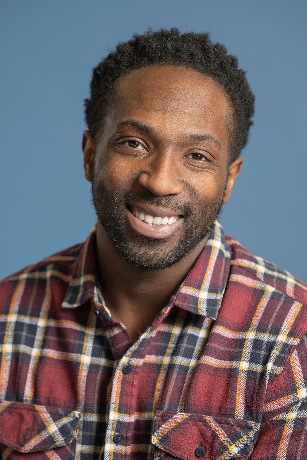 Smiling Black man with beard wearing a plaid flannel shirt against a blue background.