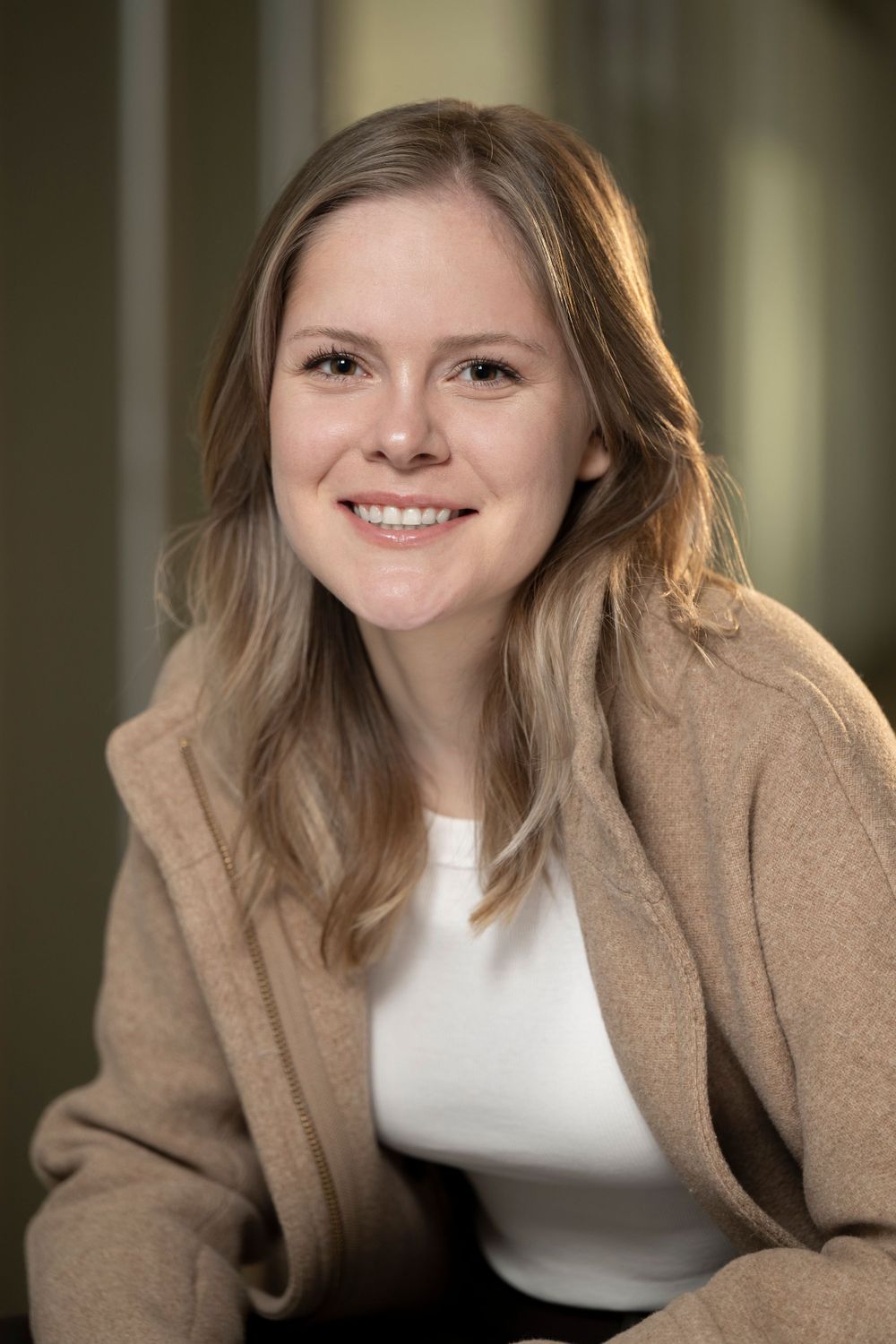 Smiling young woman with blonde hair wearing a beige coat poses for a professional photo.