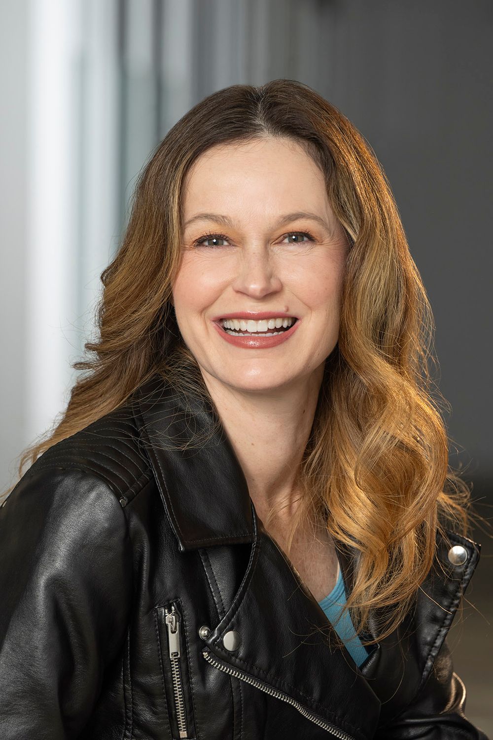 Smiling woman with auburn wavy hair wearing a black leather jacket poses indoors.
