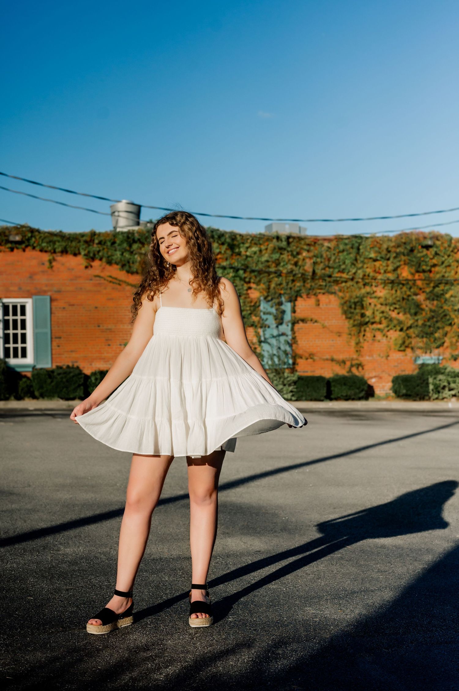 Woman in white sundress twirls in front of rustic brick building on sunny day.