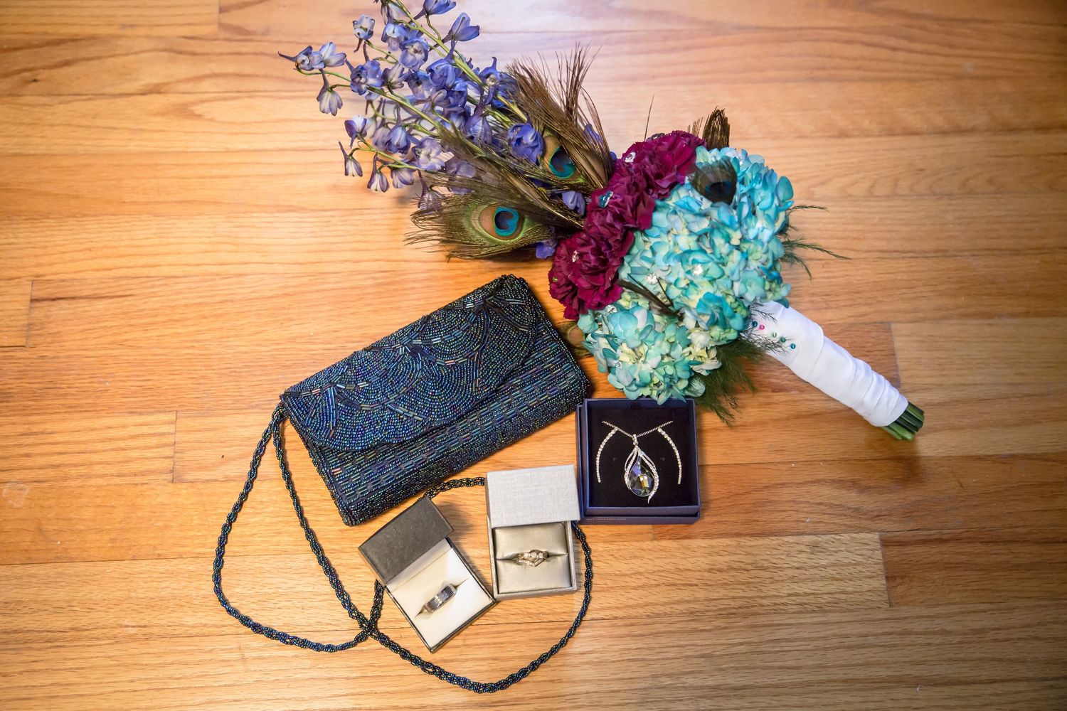 Wedding accessories including a blue bouquet, clutch, and rings arranged on wooden floor.