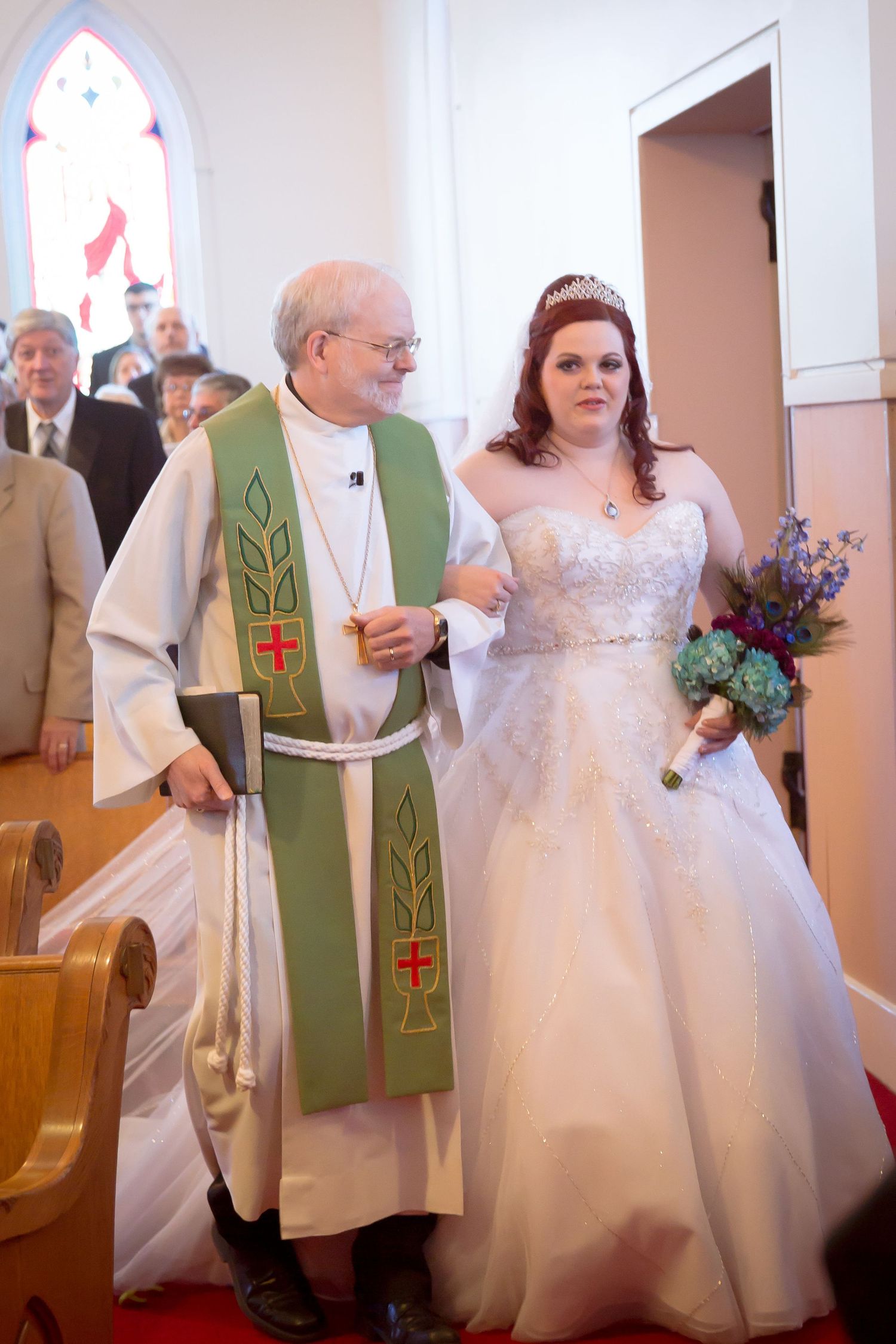 Bride in white dress walks down church aisle accompanied by minister in green ceremonial robes.