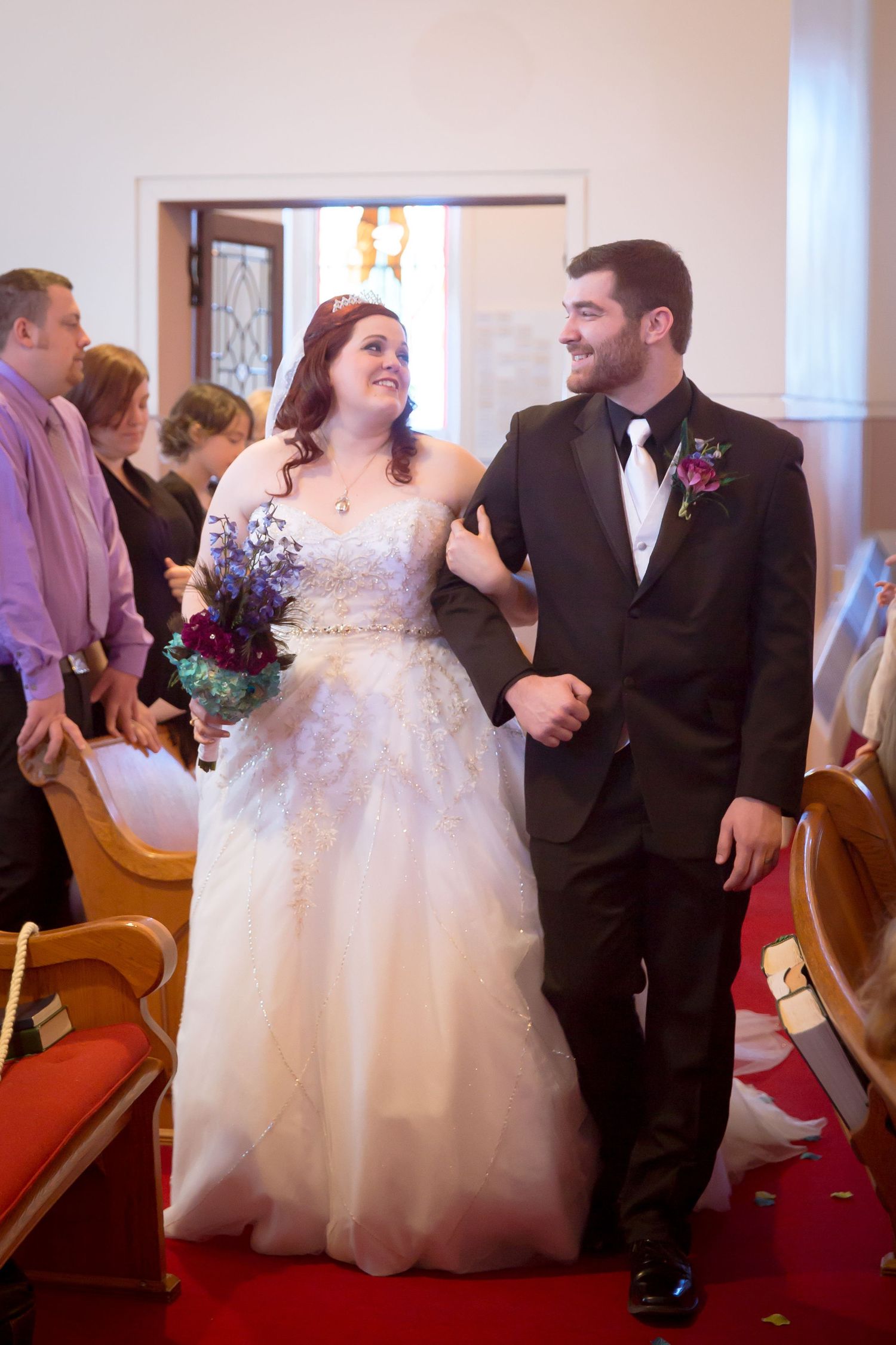 Newlyweds share joyful moment walking down church aisle with purple bouquet.