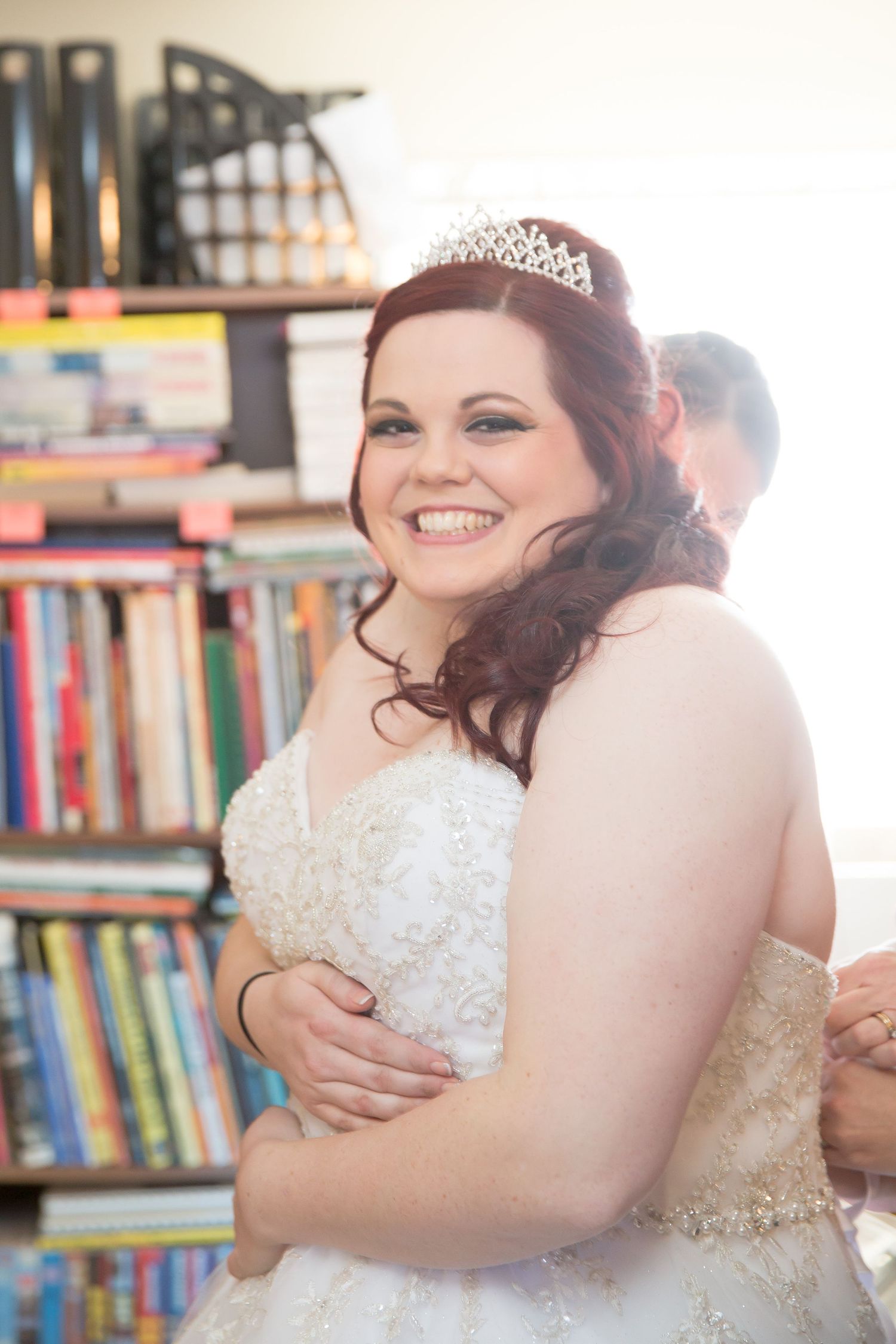 A bride in a white wedding dress and tiara smiles brightly in front of a bookshelf.