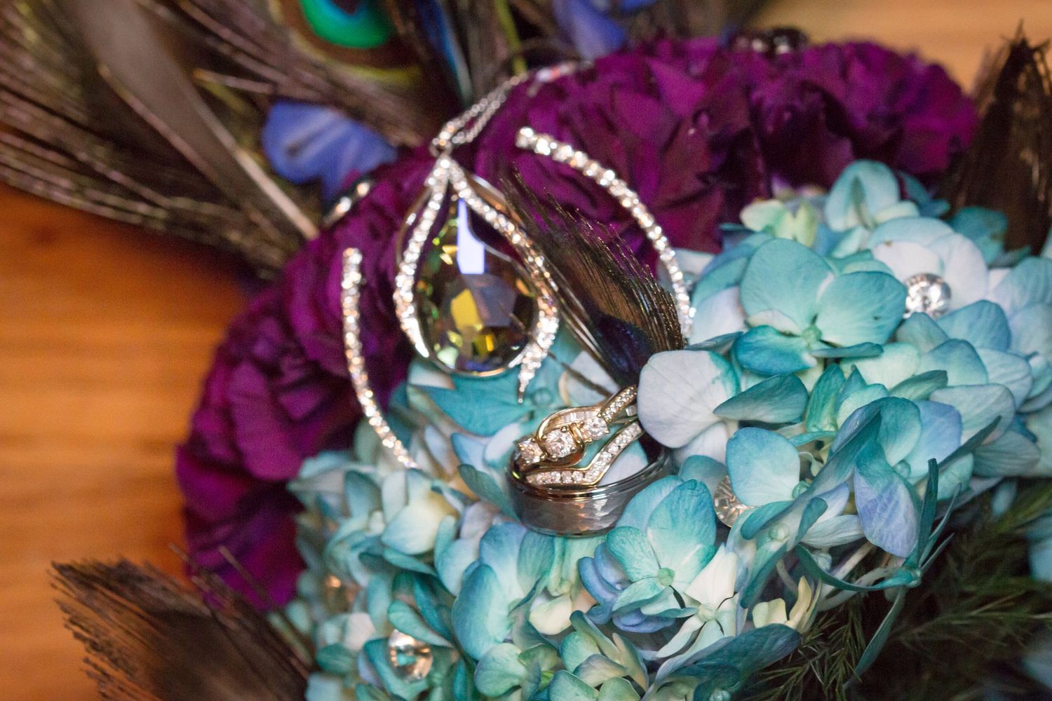 Wedding rings displayed on blue hydrangeas and purple flowers with peacock feathers.