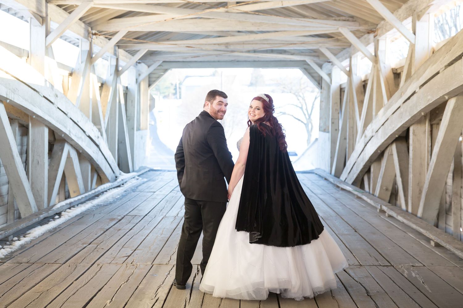 Wedding couple walking through a rustic covered bridge with the bride wearing a black cape over her dress.