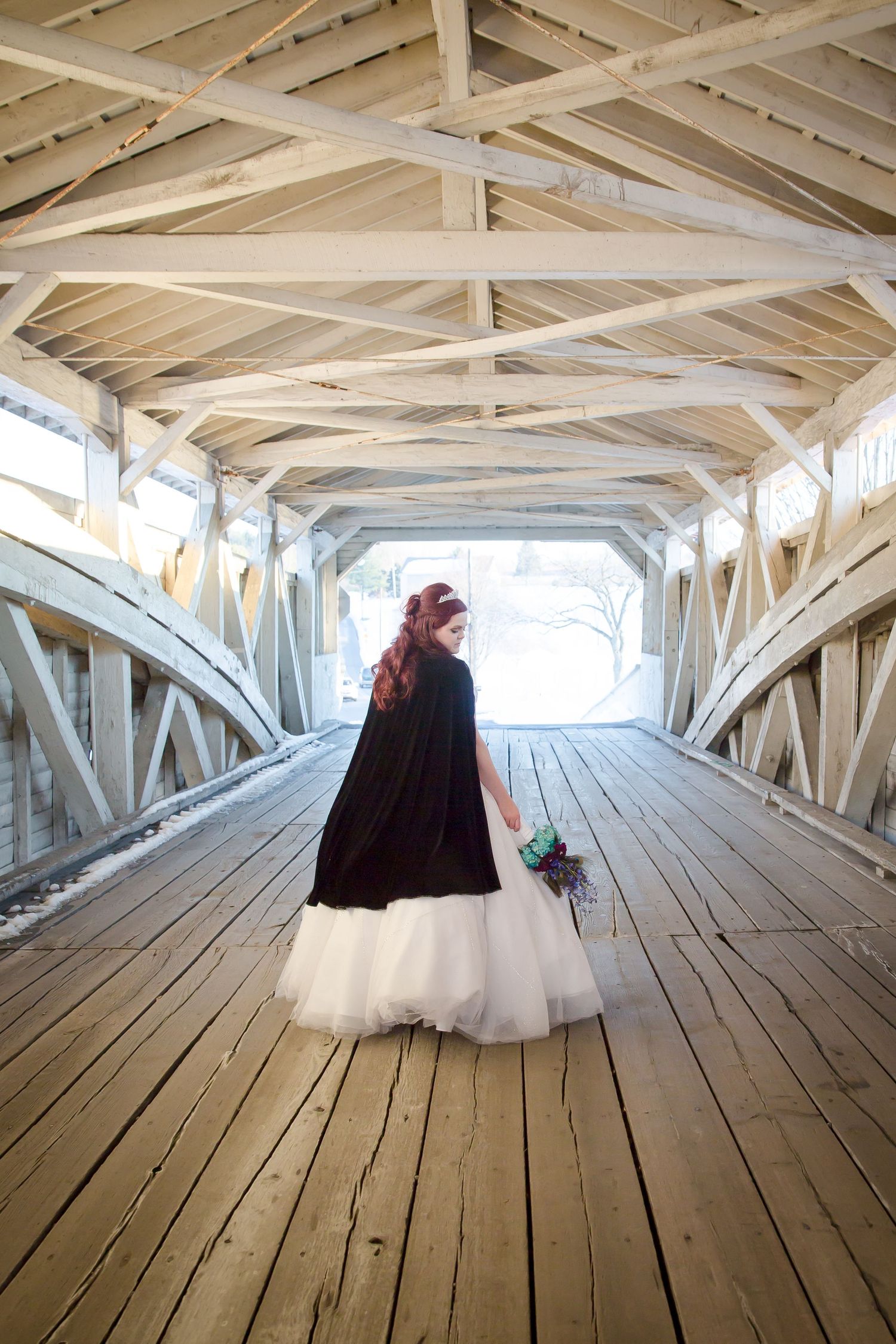 Wedding dress silhouette walking through sunlit covered bridge with rustic beams.