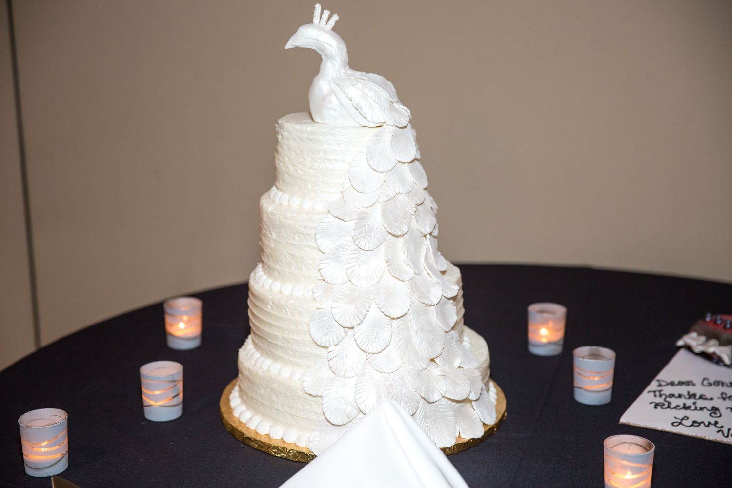 A white wedding cake decorated with peacock details and surrounded by small candles.
