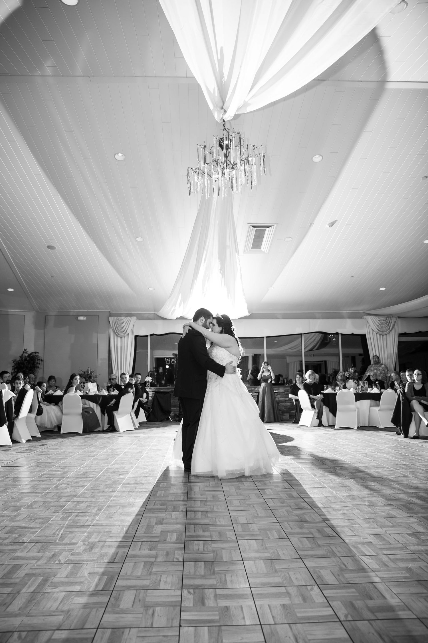 A black and white photo of a couple's first dance under an elegant chandelier and draped ceiling.