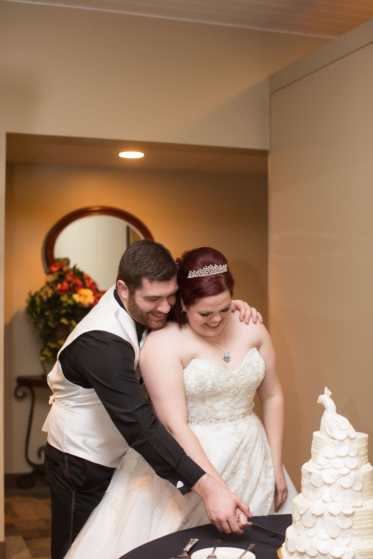 Newlyweds laugh together while cutting their tiered white wedding cake.